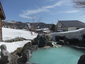 水色の濁り湯の露天風呂で、背景には山が見える。岩船の周りには、１メートルぐらいの雪の壁がある。